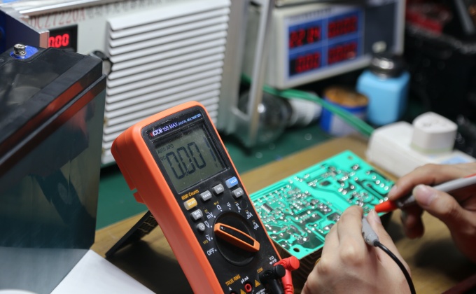 Technician inspecting battery charger PCBA electrical points with a digital multimeter
