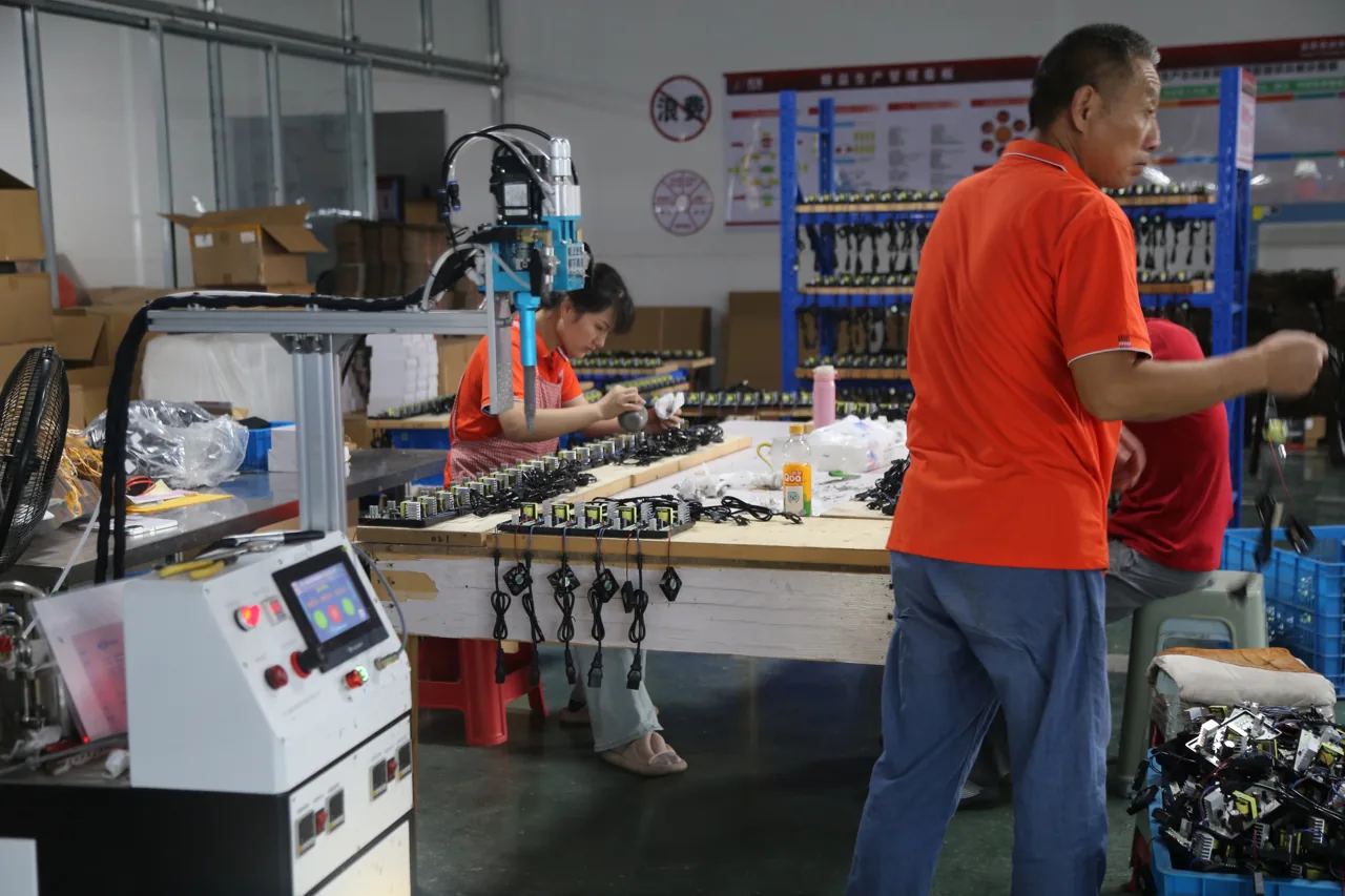 Workers handling charger assemblies and attached cables at a factory production station.