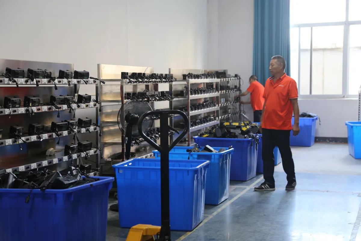 Linjia staff beside charger racks and blue production bins in the factory burn-in area.